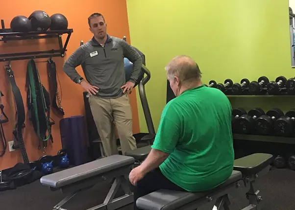 Stroke Survivor Sitting on Bench at Anytime Fitness With Personal Trainer